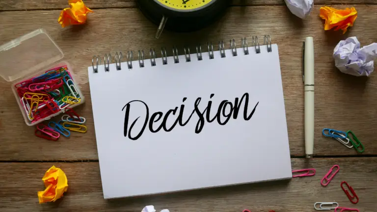 Open spiral notebook with the word 'Decision' in elegant script, surrounded by office supplies and crumpled papers on a wooden desk.