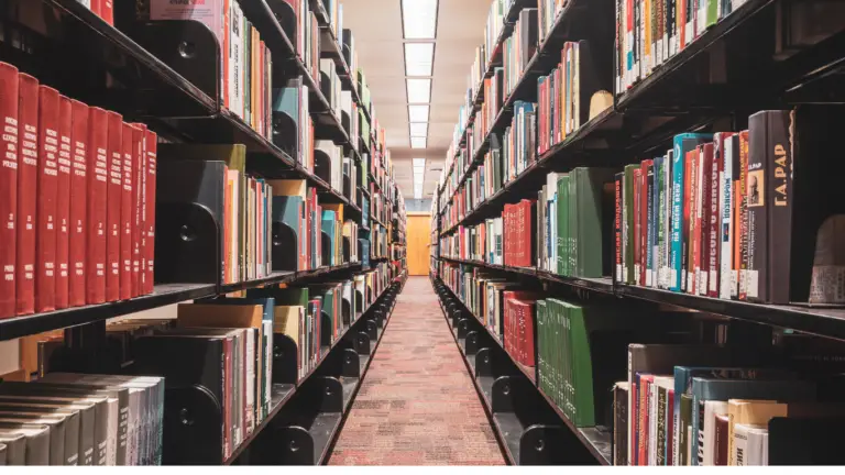 Long library aisle between tall shelves filled with colorful books, extending toward a door at the far end.
