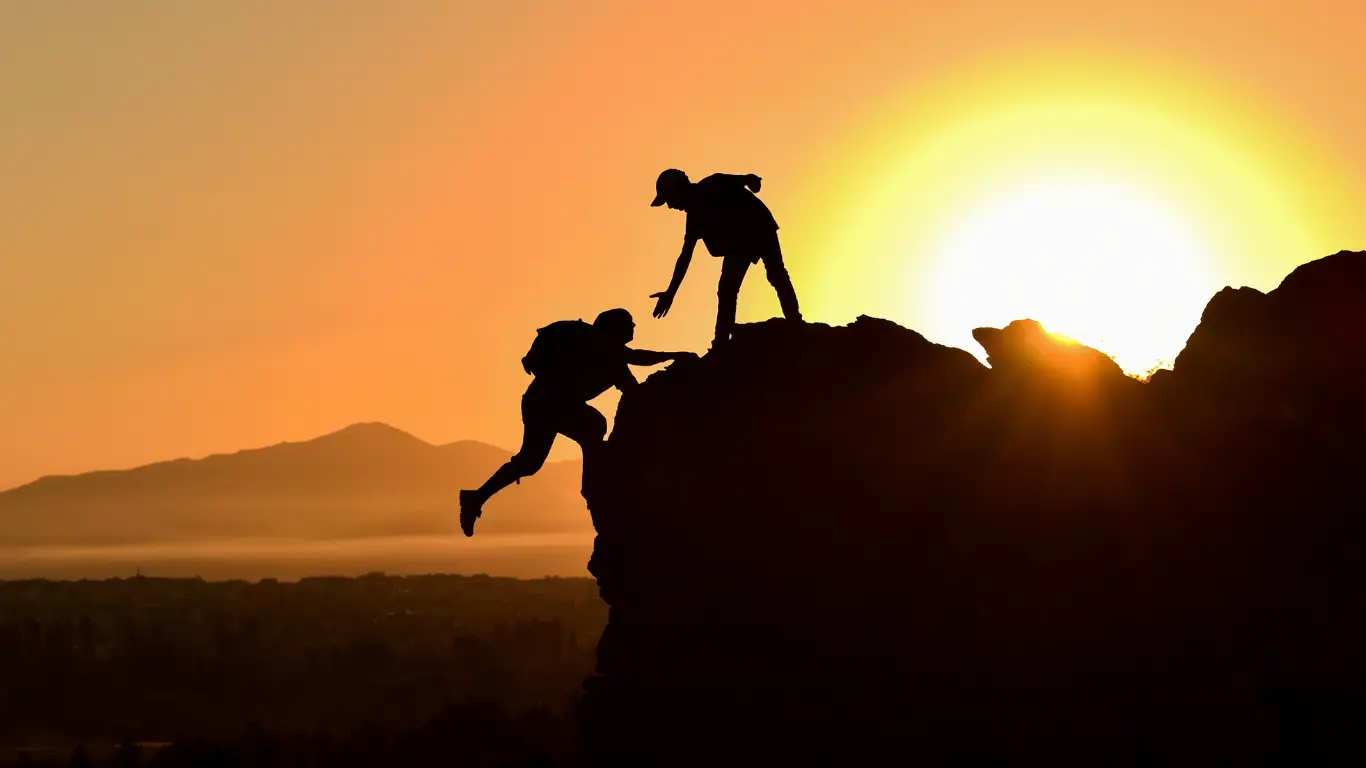 Trusts | Cybersecurity and Managed Security Services Two silhouetted climbers on a rock ledge at sunset, one reaches to help the other on the ascent.