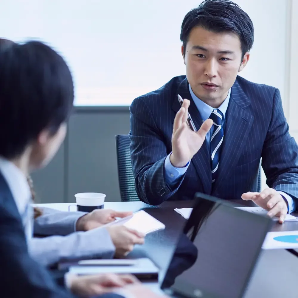 Man in a dark pinstripe suit gestures with a pen while speaking at a business meeting around a conference table with laptops and documents present.